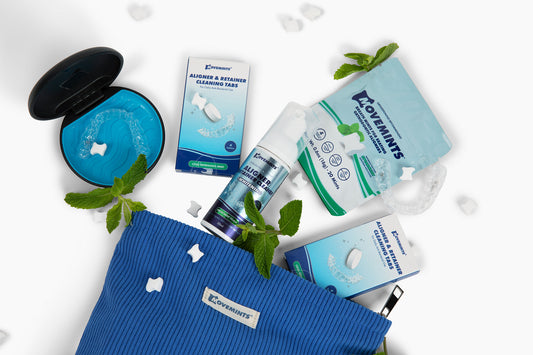 A blue corduroy zipper pouch on a white surface with Movemints cleaning tablets, whitening foam and aligner mints coming out of it. Mint leaves, loose mints and an Invisalign case also in shot